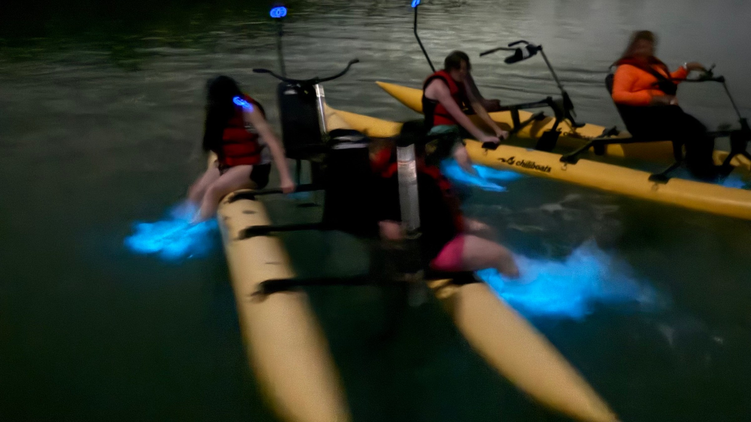 Three people on yellow water bikes with blue bioluminescent trails at night.