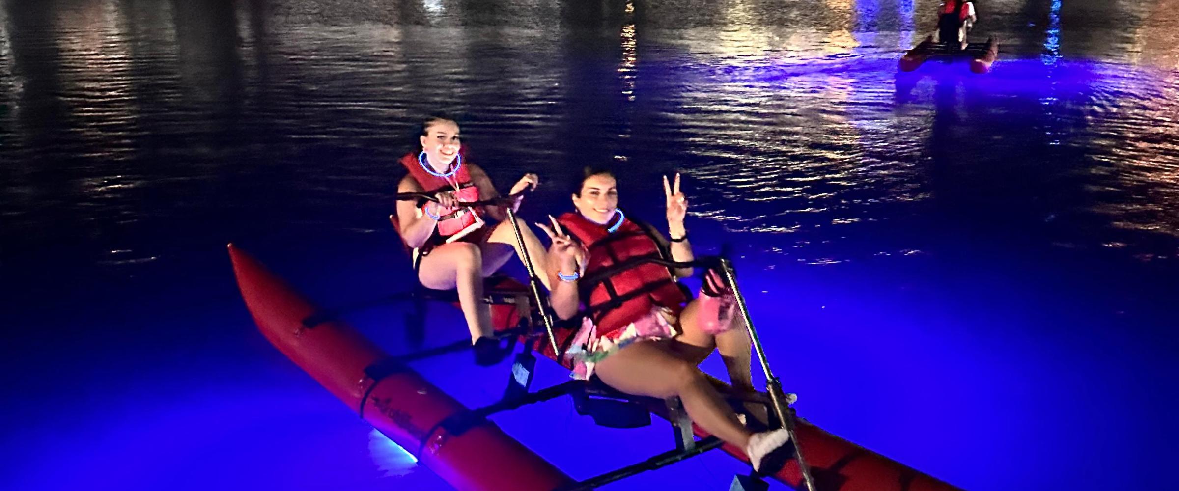 People kayaking at night with blue lights, marina and boats in the background.