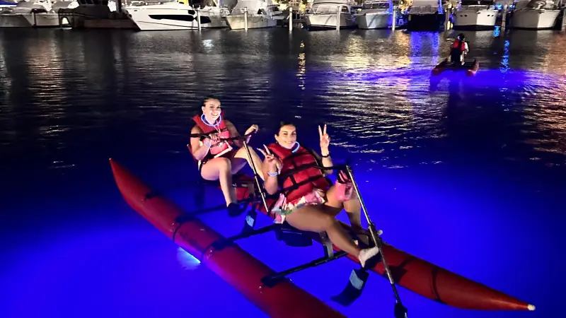 People kayaking at night with blue lights, marina and boats in the background.
