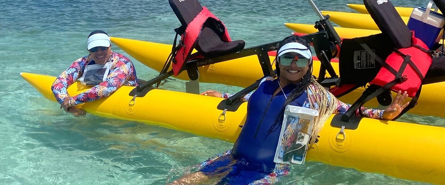 Two people in colorful outfits relaxing on yellow water bicycles in clear blue water.