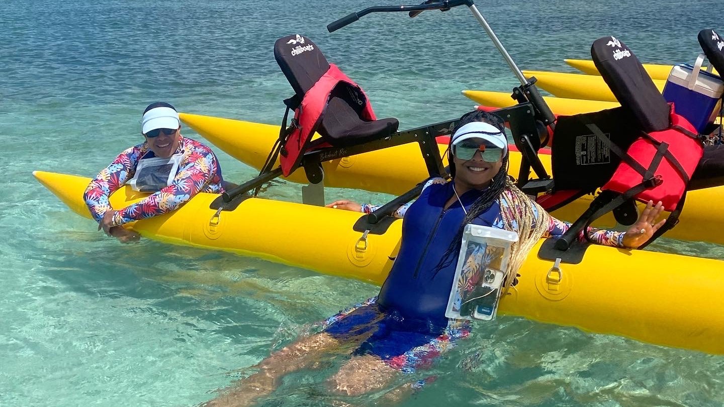 Two people in colorful outfits relaxing on yellow water bicycles in clear blue water.