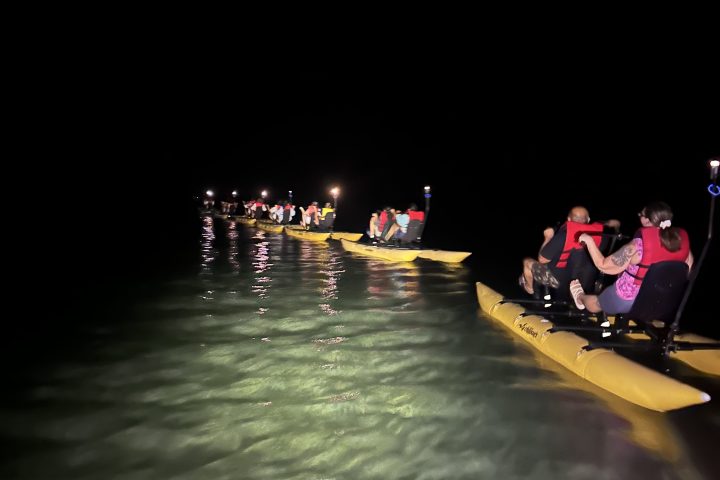 Nighttime kayaking tour with people on illuminated yellow kayaks over calm water.
