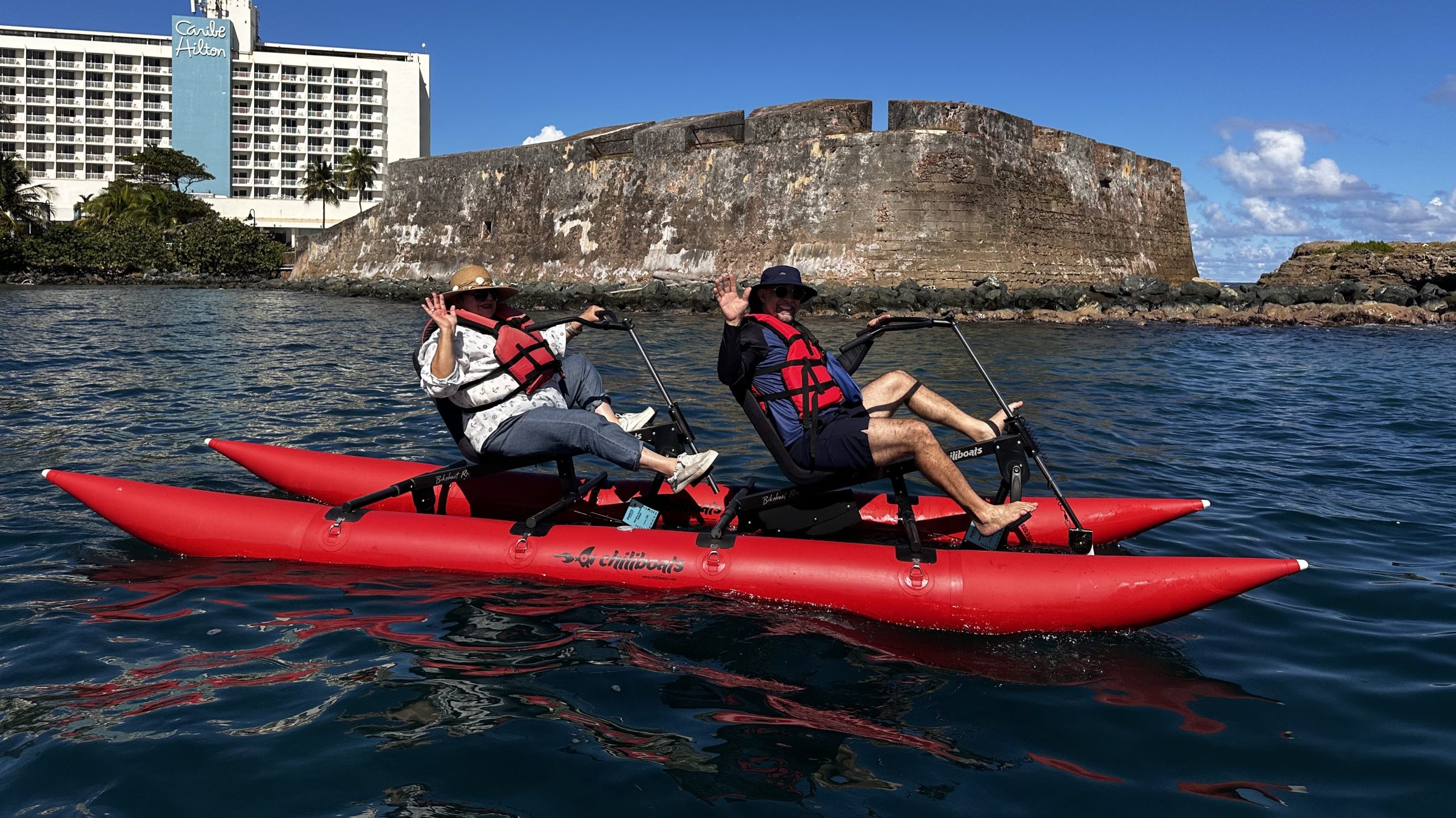 Two people in red pedal kayaks wave in front of a historic fort and hotel by the sea.