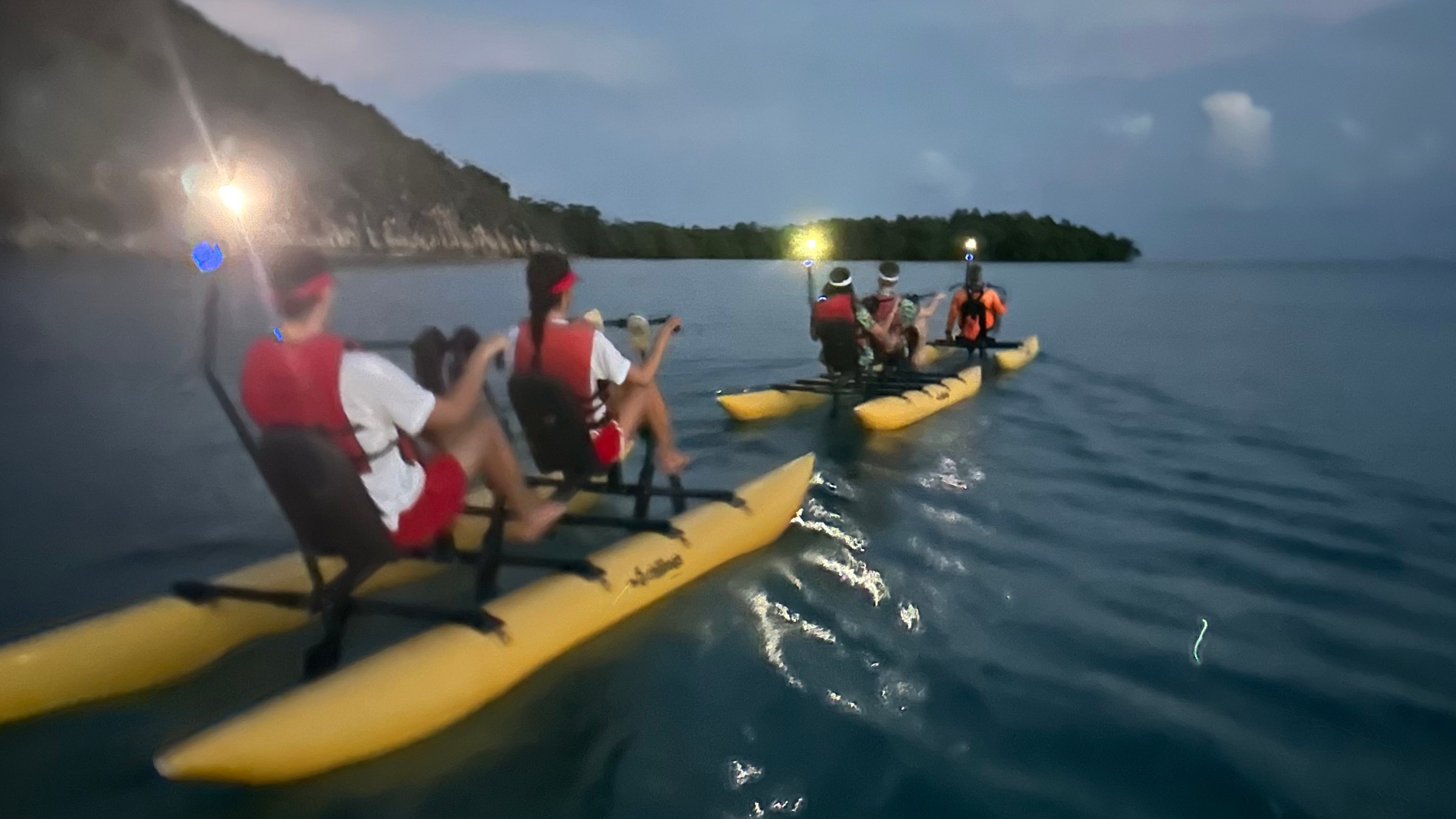 Group kayaking on a calm sea at dusk with headlamps shining, island visible in background.