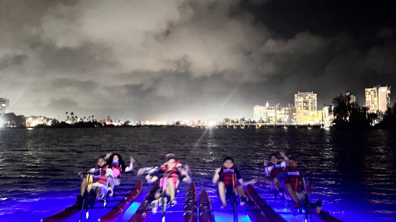 Group paddle boating on illuminated water at night with city lights in background.