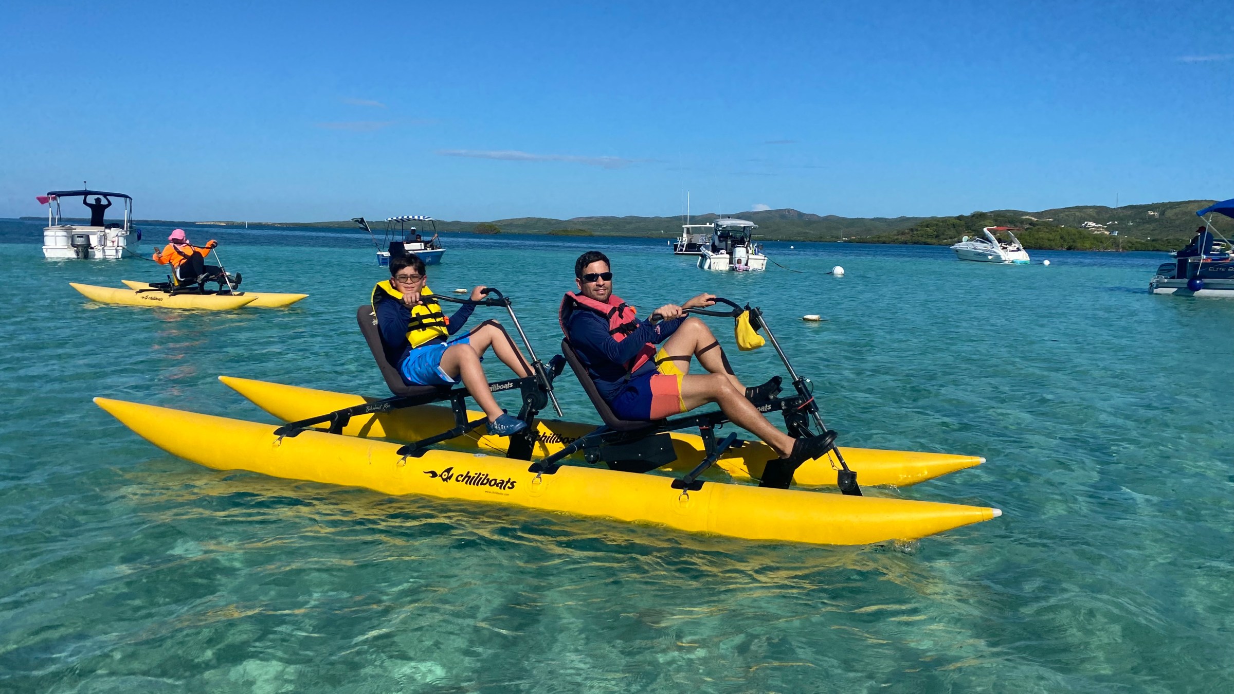Two people ride a yellow water bike on clear blue water, surrounded by boats and distant green hills.