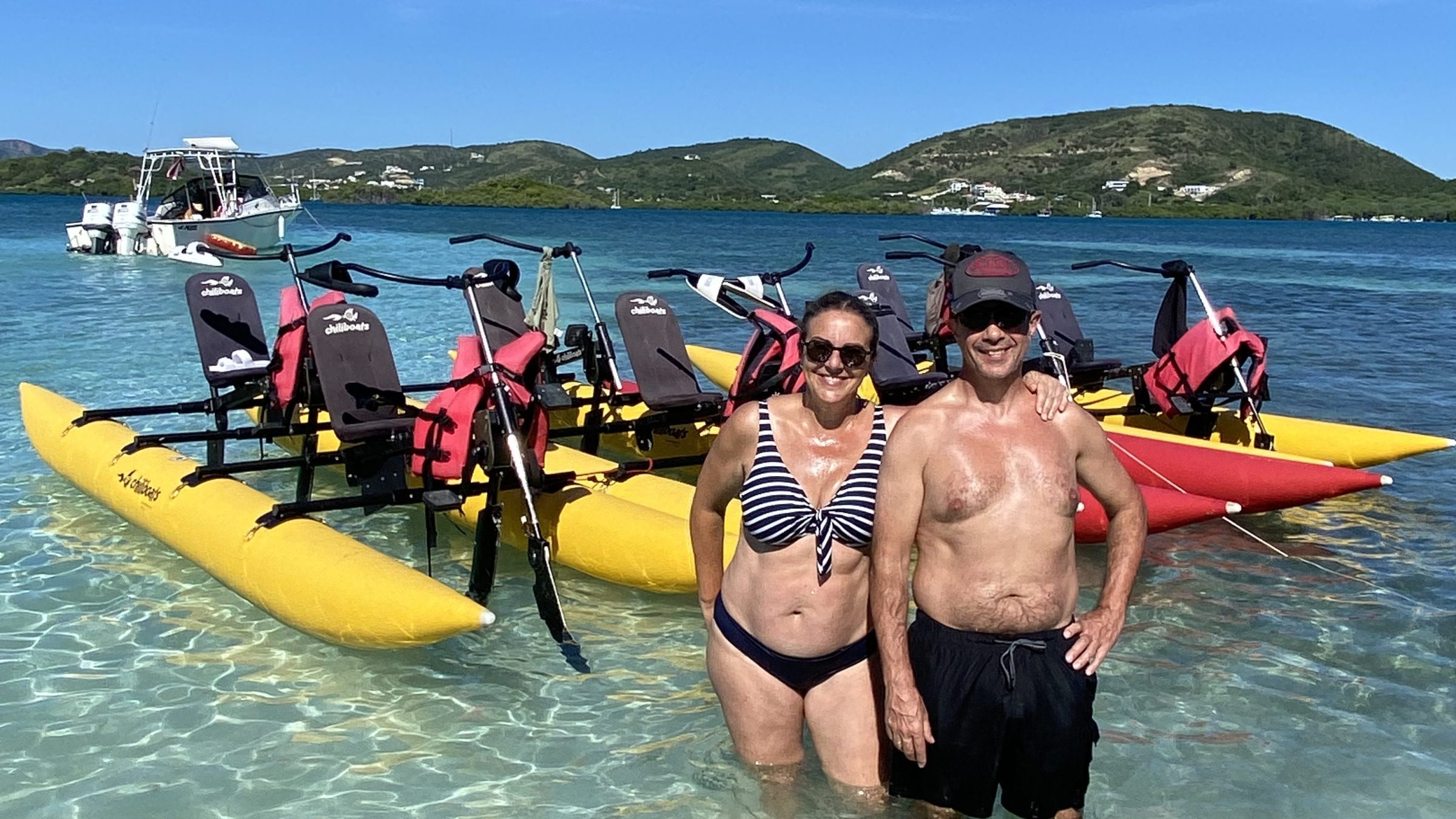 Two people stand in shallow water near yellow water bikes, with green hills and a blue sky in the background.