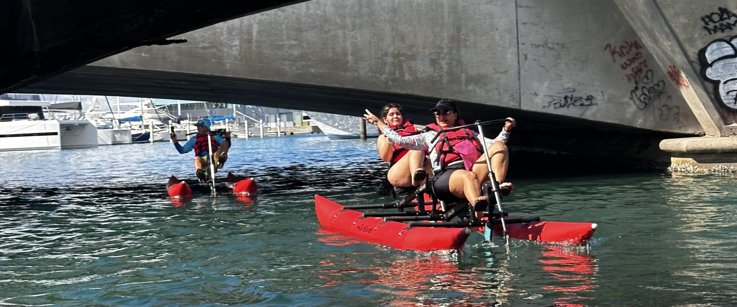 People kayaking under a concrete bridge over calm water.