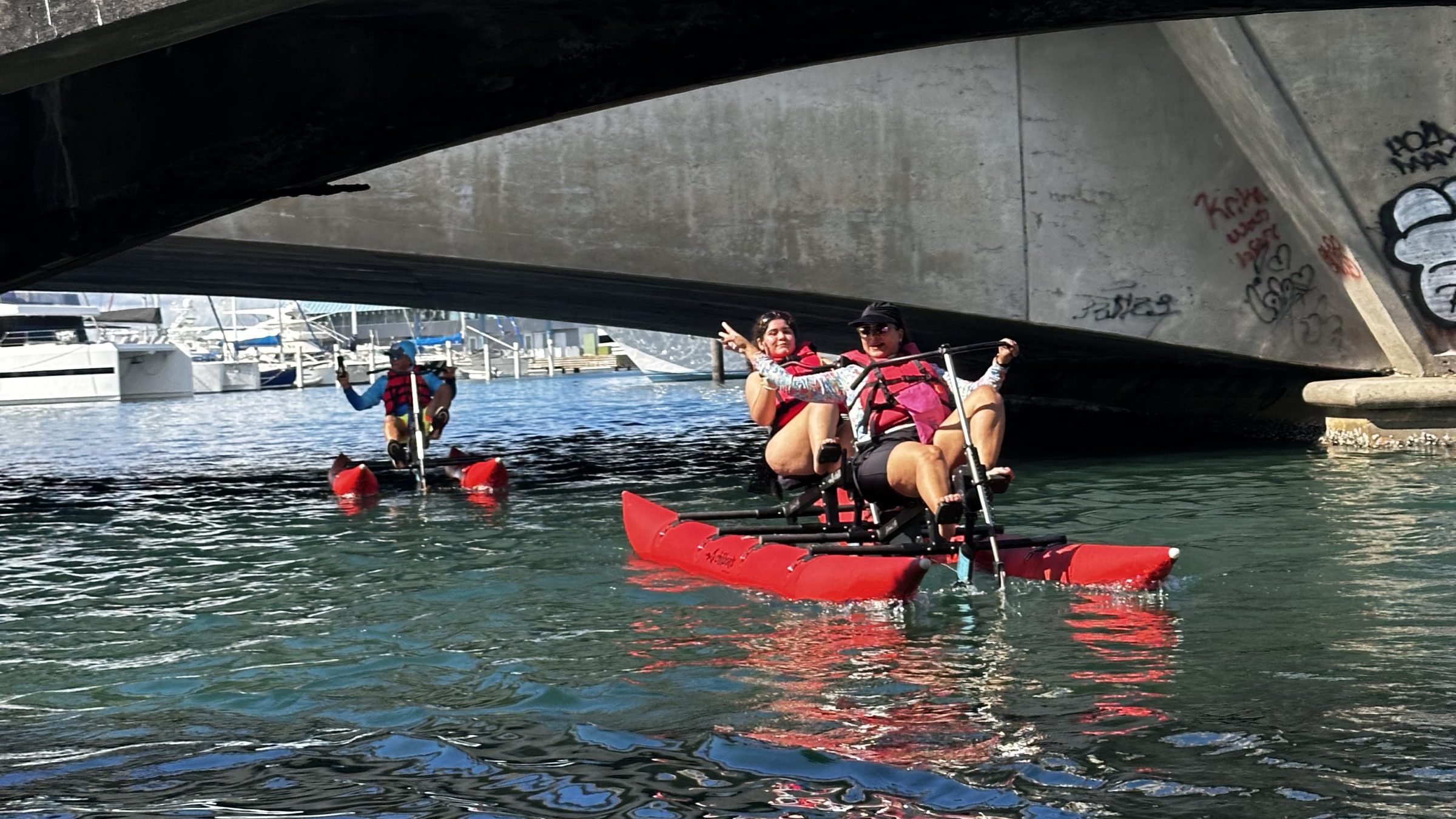 People kayaking under a concrete bridge over calm water.