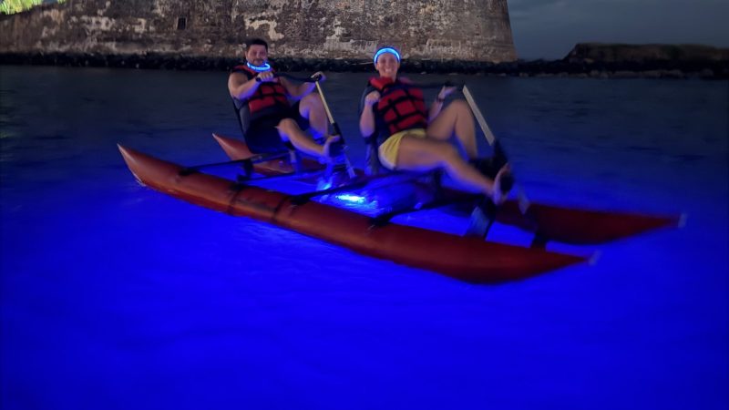 Two people kayaking at night with a fort in the background, water glowing blue.