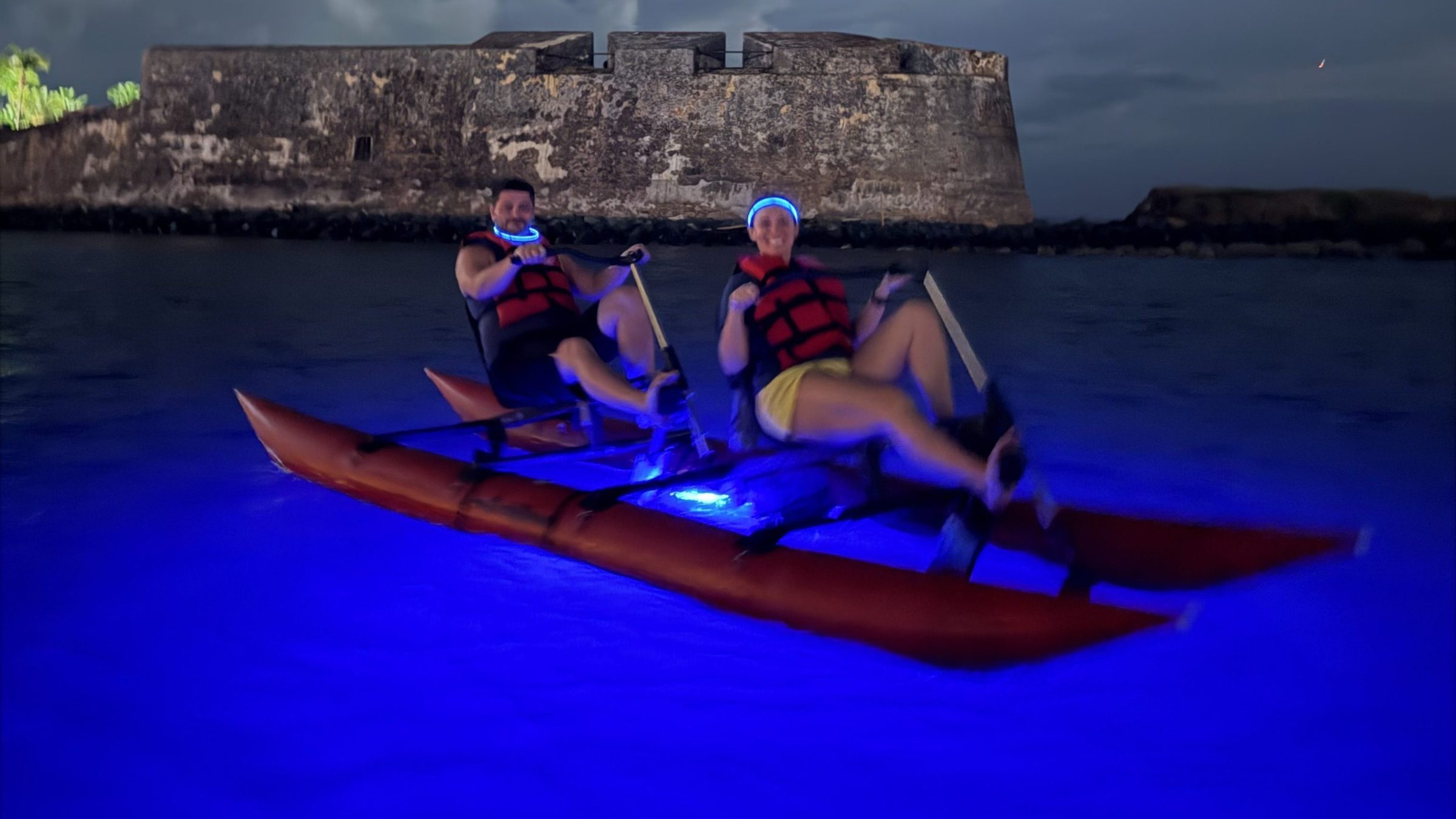 Two people kayaking at night with a fort in the background, water glowing blue.