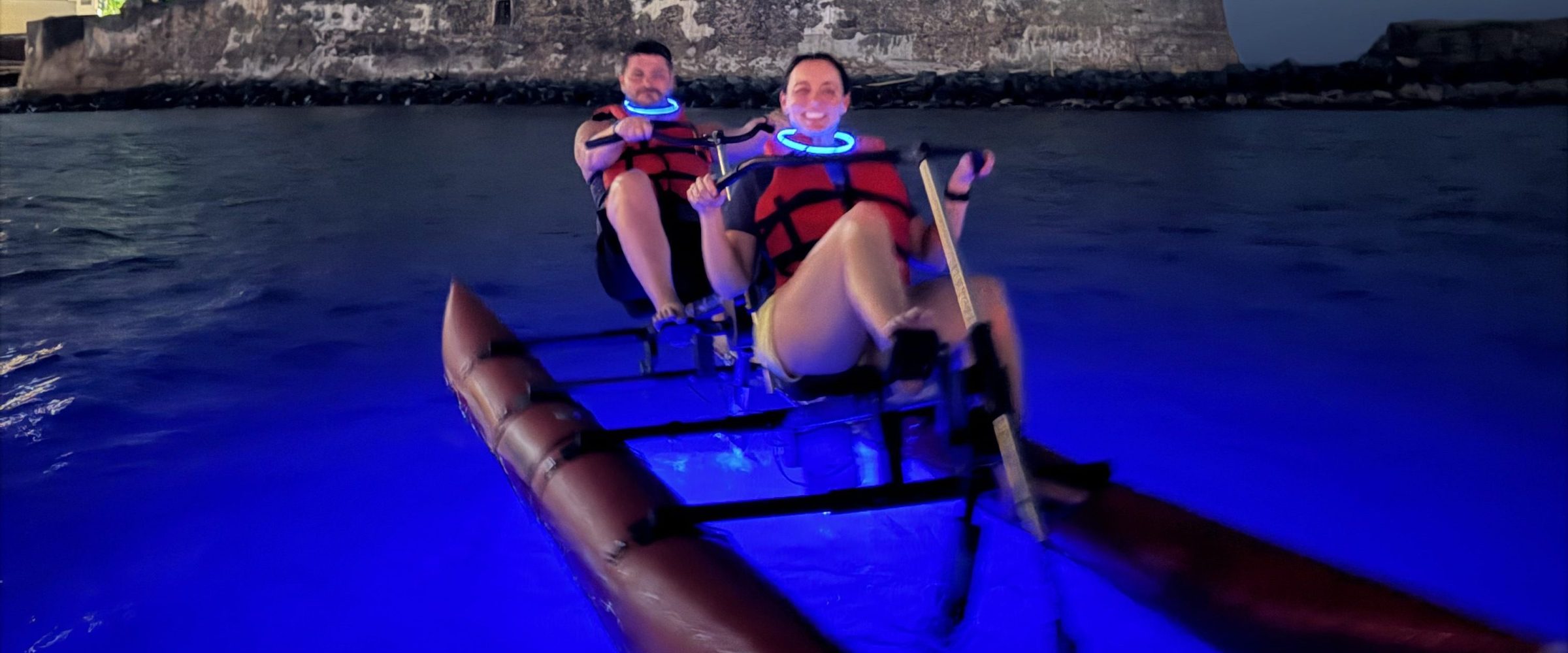Two people kayaking on illuminated water near a fortress at night.