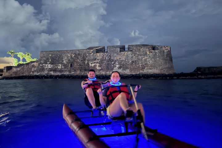 Two people kayaking at night on blue-lit water near a historic fort.
