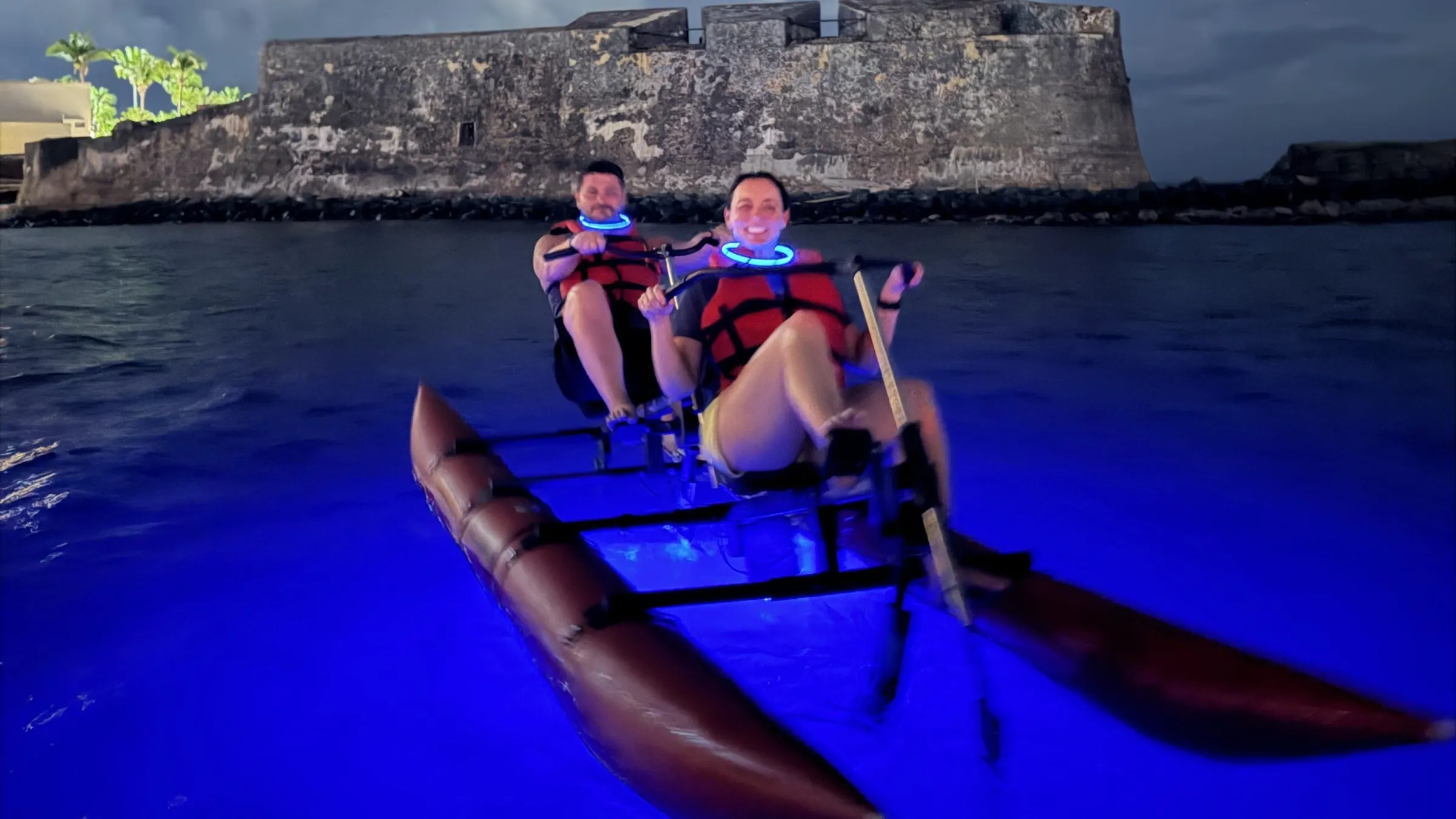 Two people in life vests paddling a canoe in blue-lit water at night near a stone fortress.