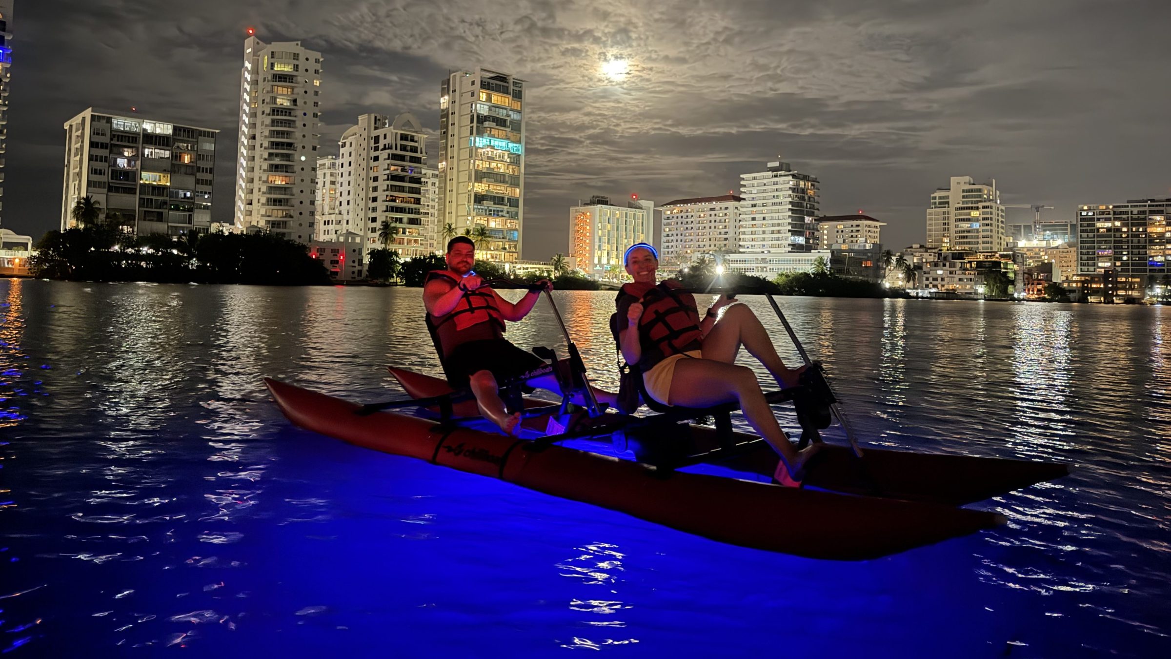 Two people kayaking at night under a cloudy sky with city skyline in the background.