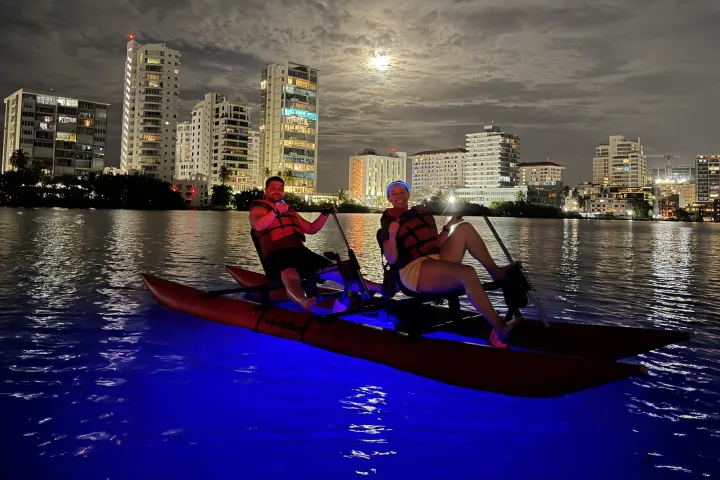 Two people kayaking at night under a cloudy sky with city skyline in the background.