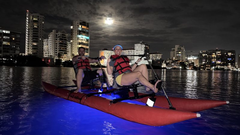 Two people in life vests kayaking on a lit-up night river with city skyline in background.