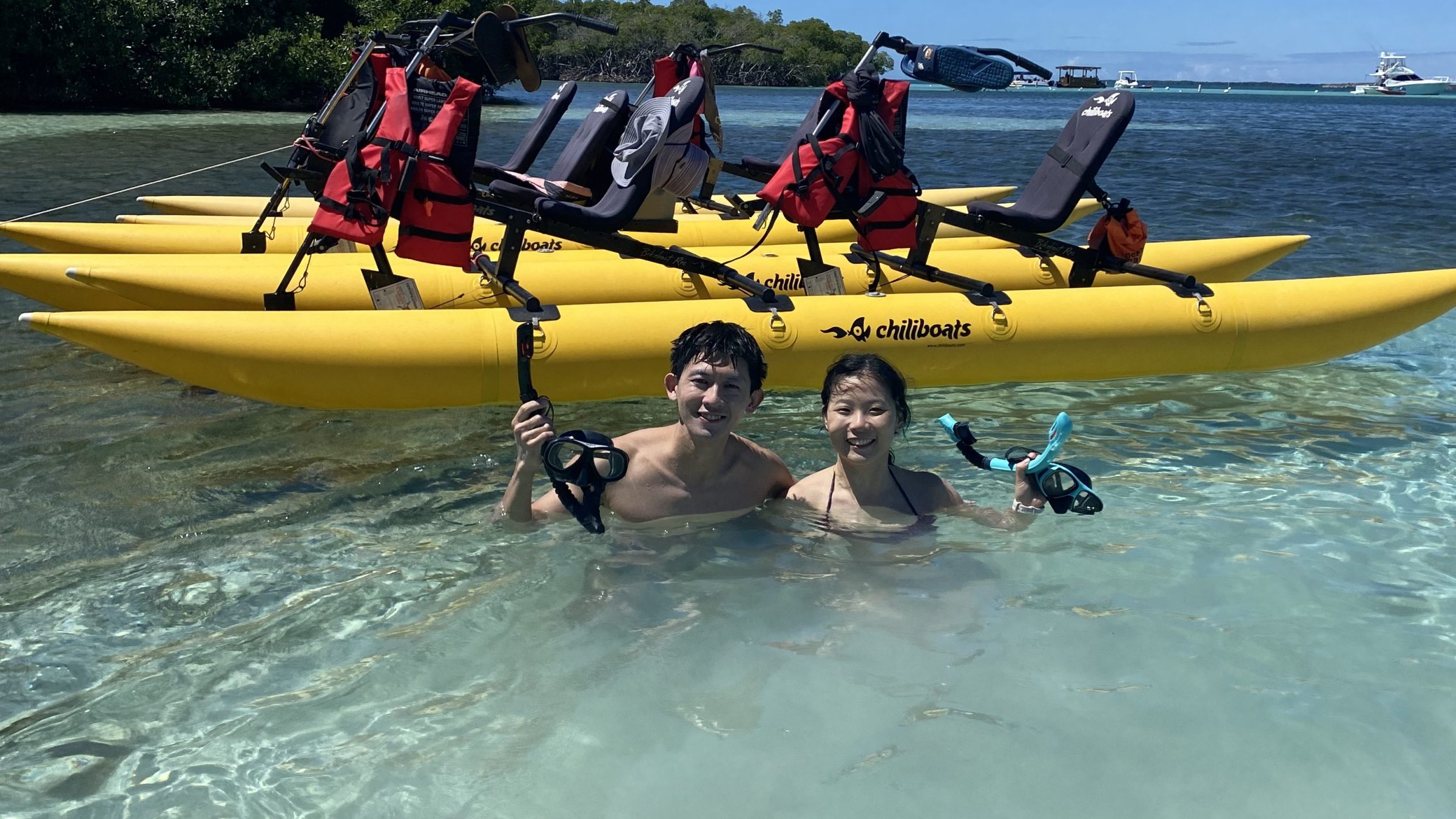 Two people in water near yellow pedal boats with red life jackets, under a clear blue sky.