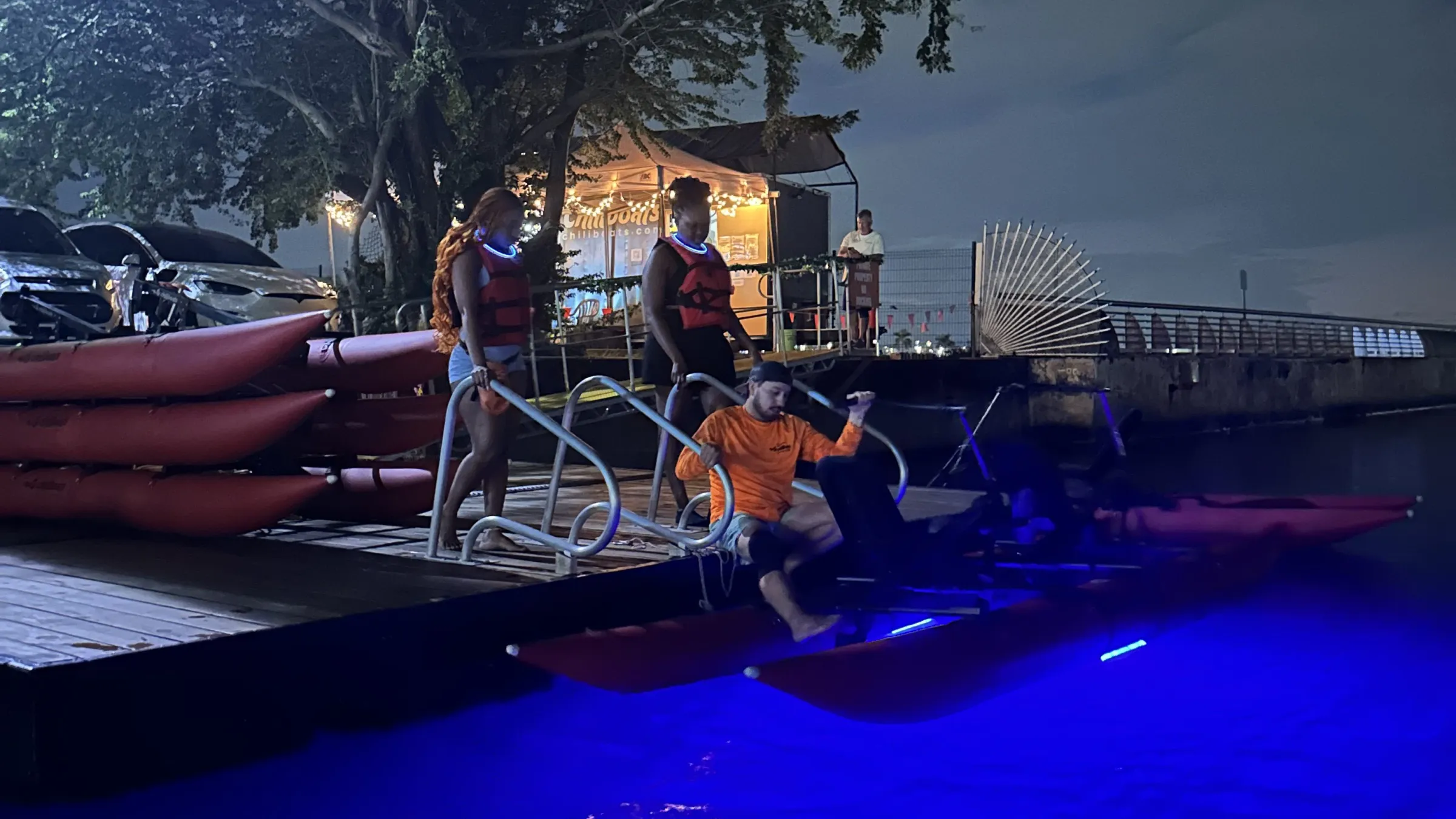 People on a dock at night with glowing blue water, kayaks, and a lit-up tent under a large tree.