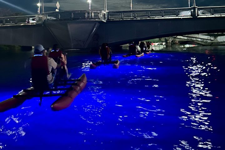 Kayakers with colorful lights paddle under a bridge at night with a cloudy moonlit sky.
