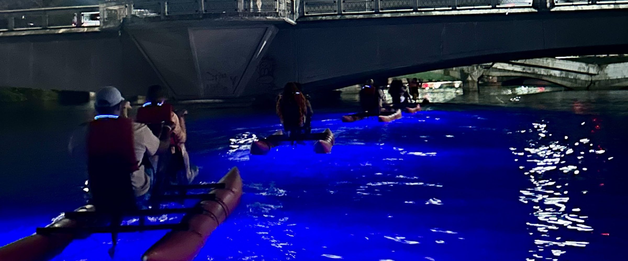 Kayakers with glowing blue lights under a cloudy night sky beneath a bridge.