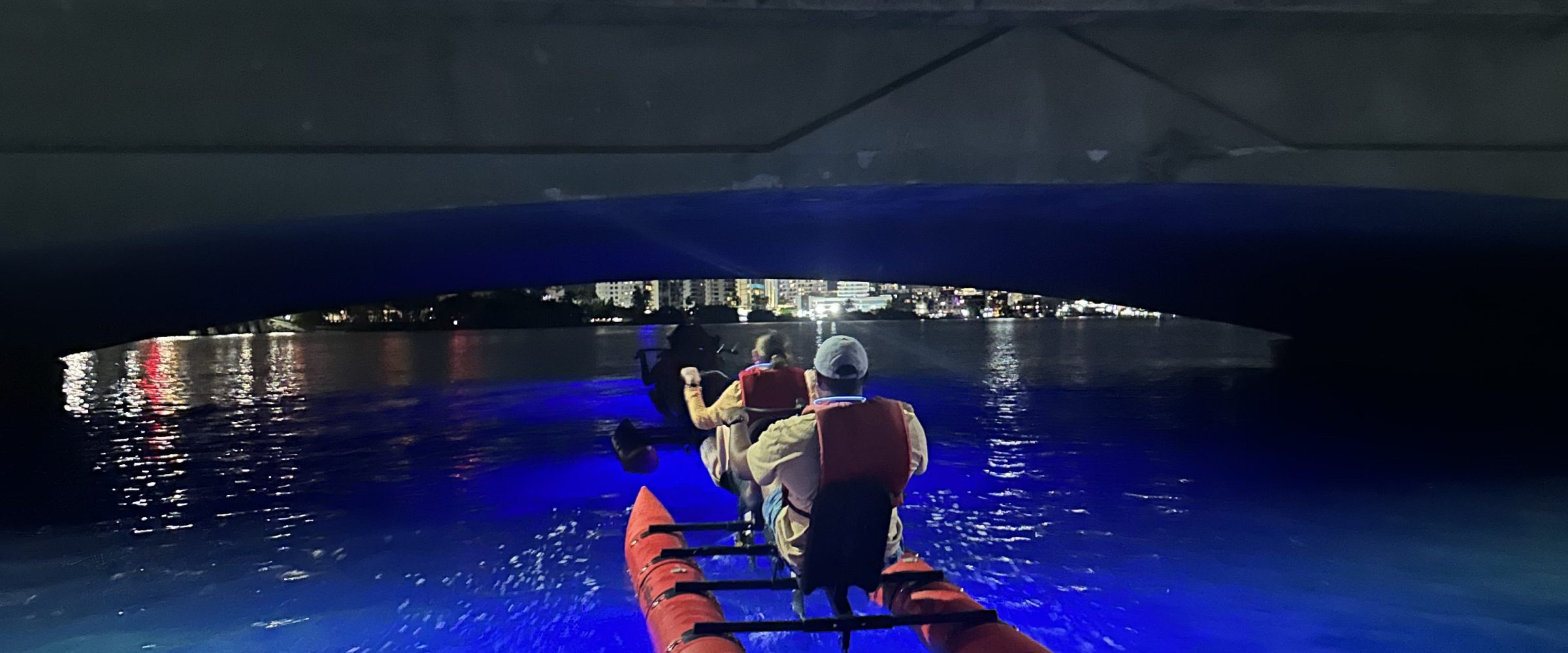 People kayaking under a bridge at night, with bright moon and blue-lit water.