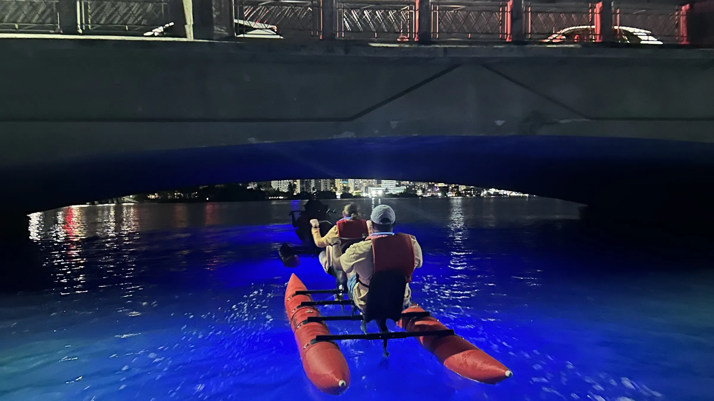 People kayaking under a bridge at night, with bright moon and blue-lit water.