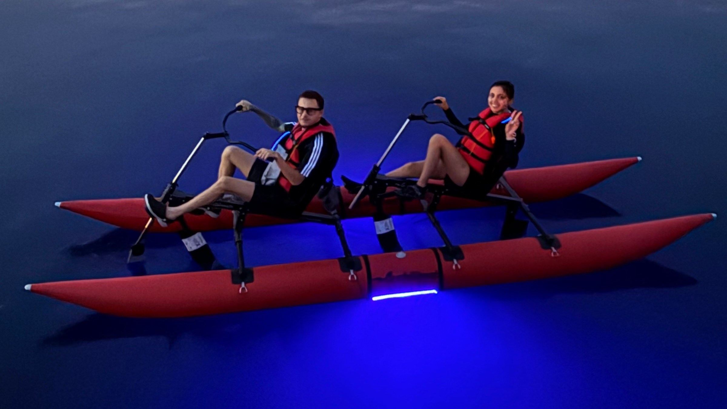 Two people in red life vests on a pedal boat with blue lights, dock and boats in background.