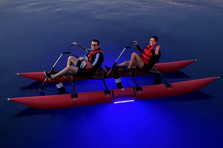 Two people in red life vests on a pedal boat with blue lights, dock and boats in background.