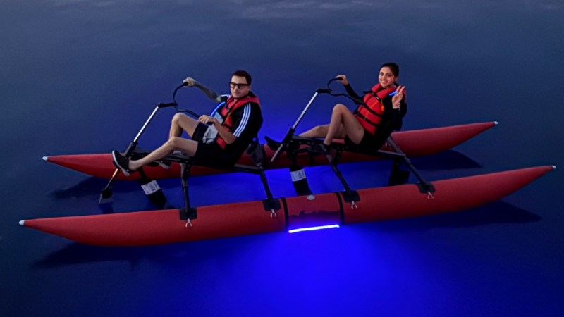 Two people in red life vests on a pedal boat with blue lights, dock and boats in background.