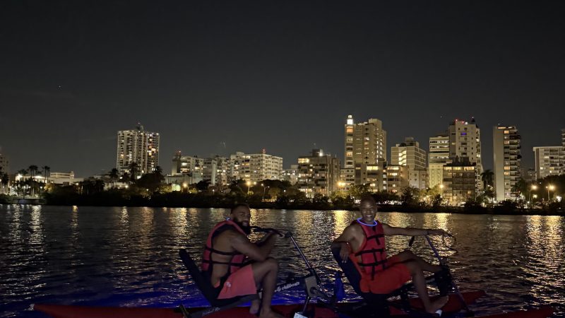 Two people on a pedal boat with blue lights on water, city buildings in the background at night.