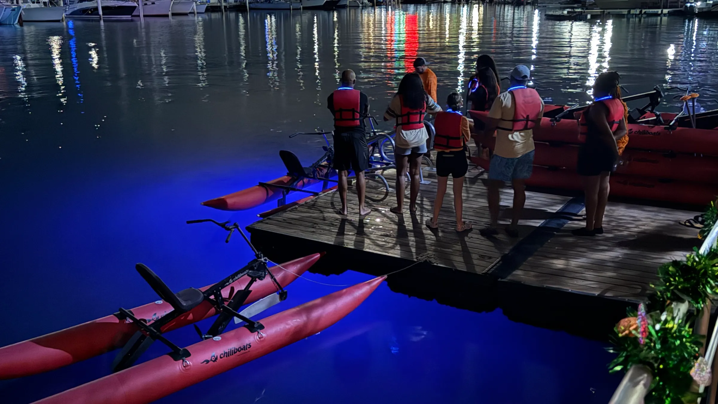 People with life jackets stand on a dock by illuminated water bikes at night.