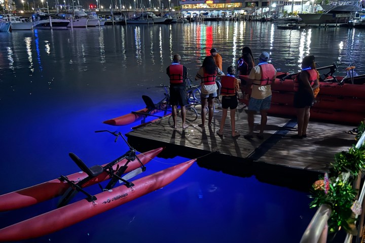 People with life jackets stand on a dock by illuminated water bikes at night.