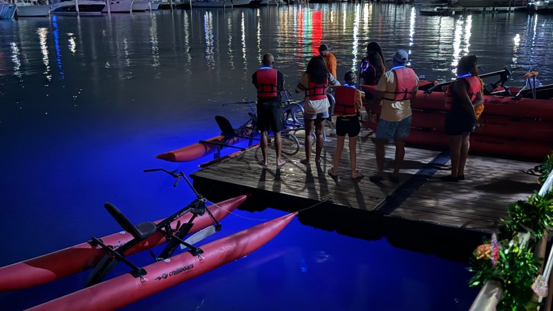 People with life jackets stand on a dock by illuminated water bikes at night.