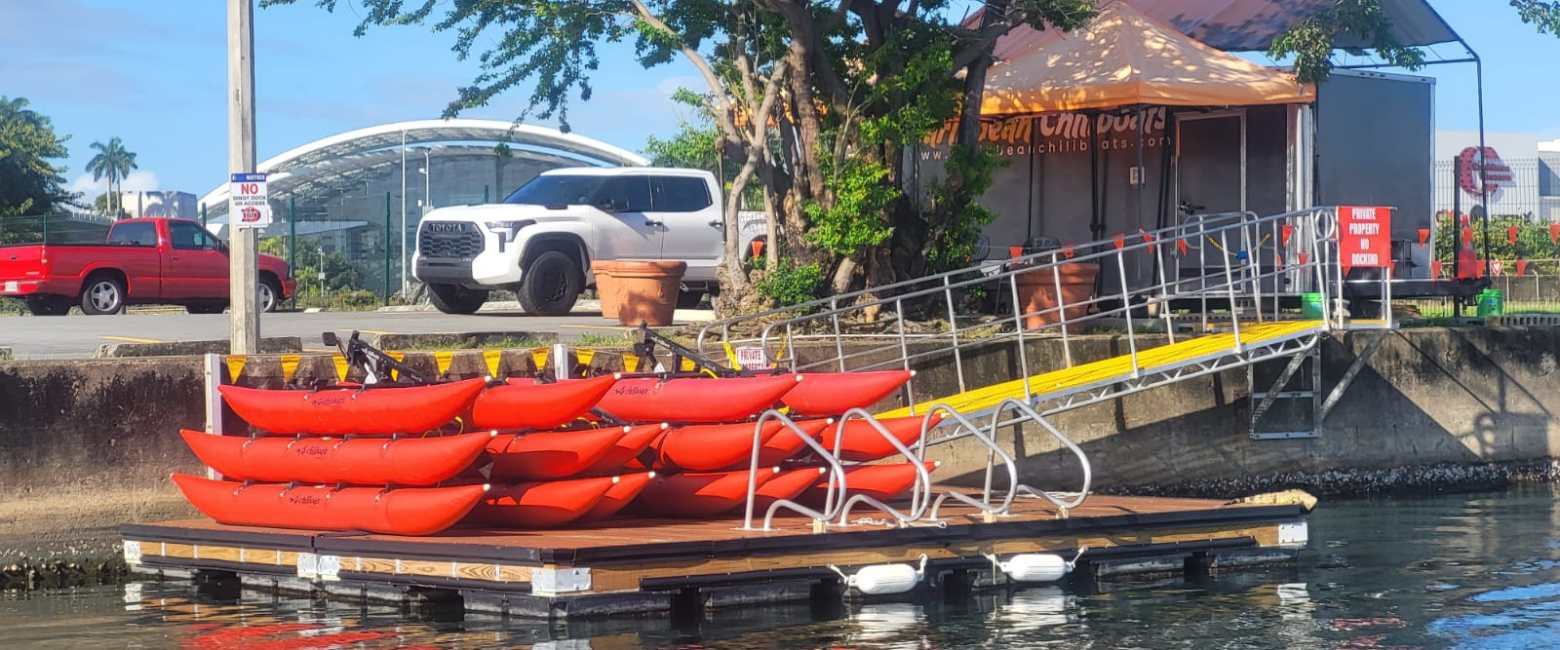 Stacked red kayaks on a dock near a ramp, with cars and trees in the background.