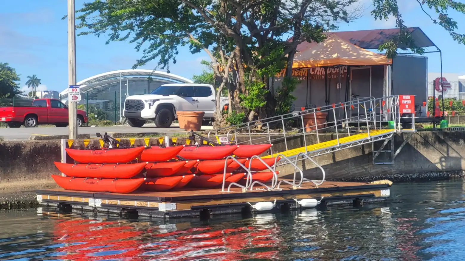 Stacked red kayaks on a dock near a ramp, with cars and trees in the background.