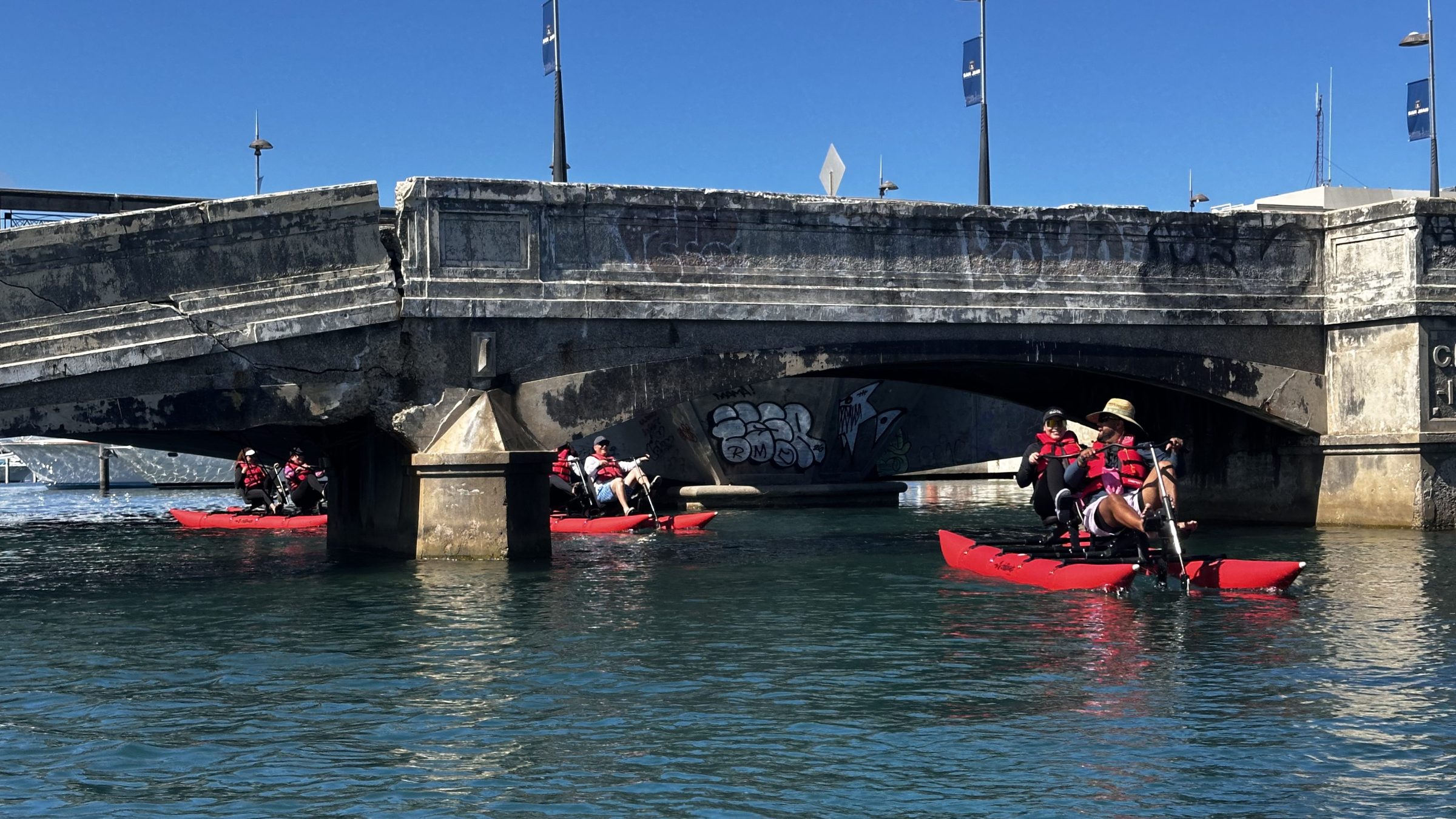 People in red kayaks paddle under a graffitied bridge on a sunny day.