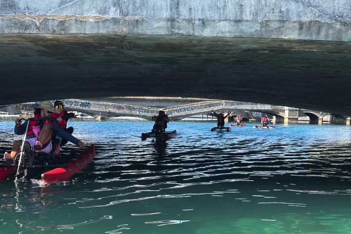 People on pedal boats pass under a graffiti-covered bridge on a sunny day.