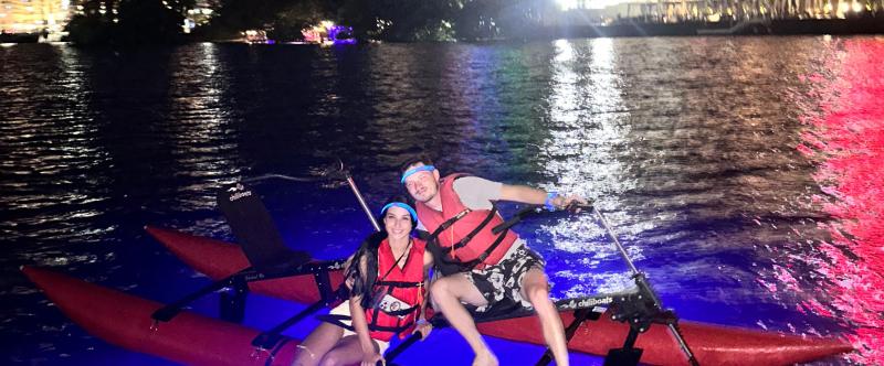 Two people on a glowing paddle board at night with city lights in the background.