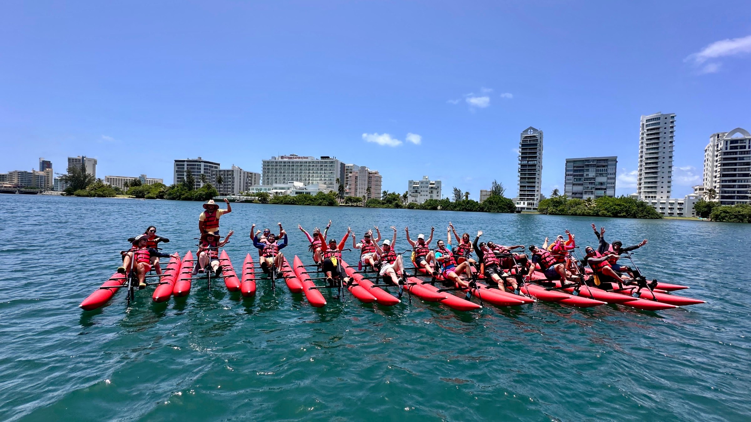 Group of people on pedal kayaks raise hands in water, city skyline in background, sunny day.