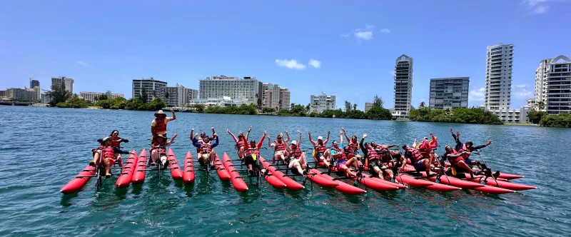 Group of people on pedal kayaks raise hands in water, city skyline in background, sunny day.