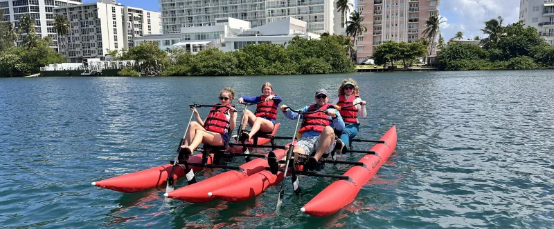 Four people on a pedal-powered watercraft with red pontoons in a sunny urban waterfront setting.