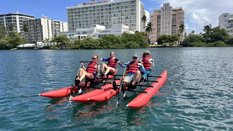 Four people on a pedal-powered watercraft with red pontoons in a sunny urban waterfront setting.