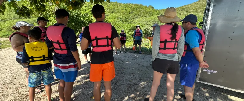 People wearing life jackets gather outdoors under a tree.