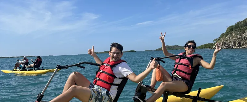Two people on yellow water bikes, wearing life jackets, giving thumbs up in a sunny, blue ocean setting.