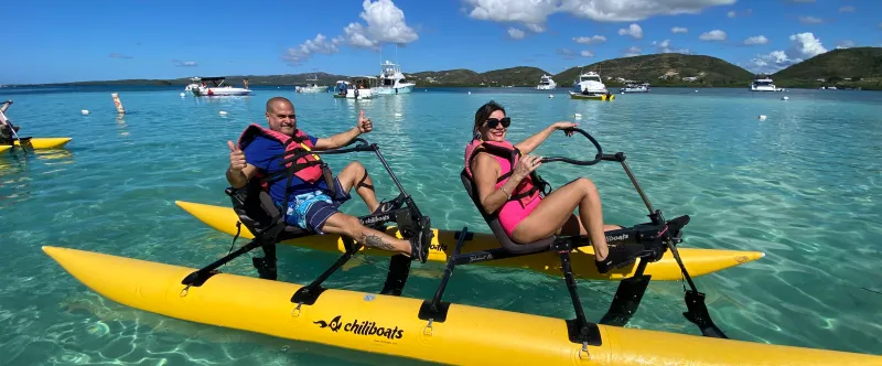 Two people on a yellow water bike in clear blue ocean with boats and hills in the background.