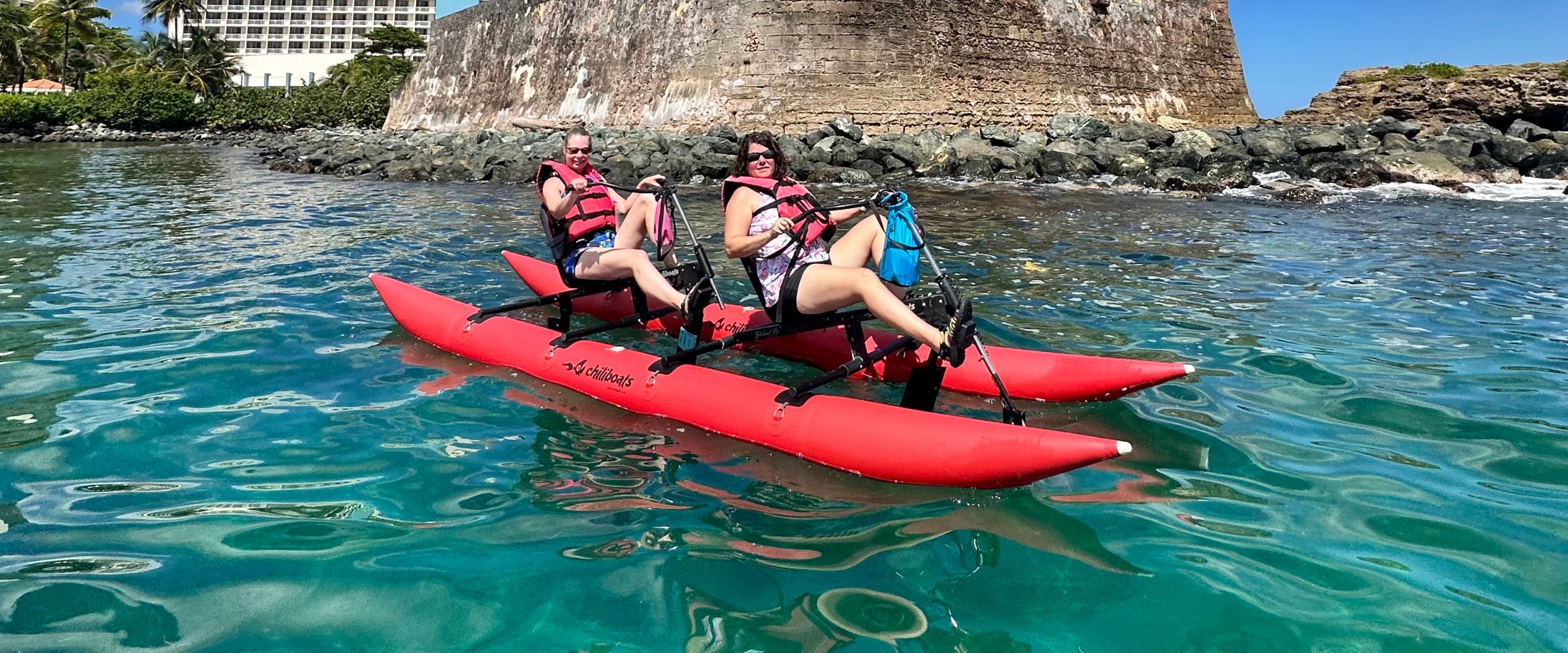 Two people ride a red waterbike on clear blue water near a historic fort on a sunny day.