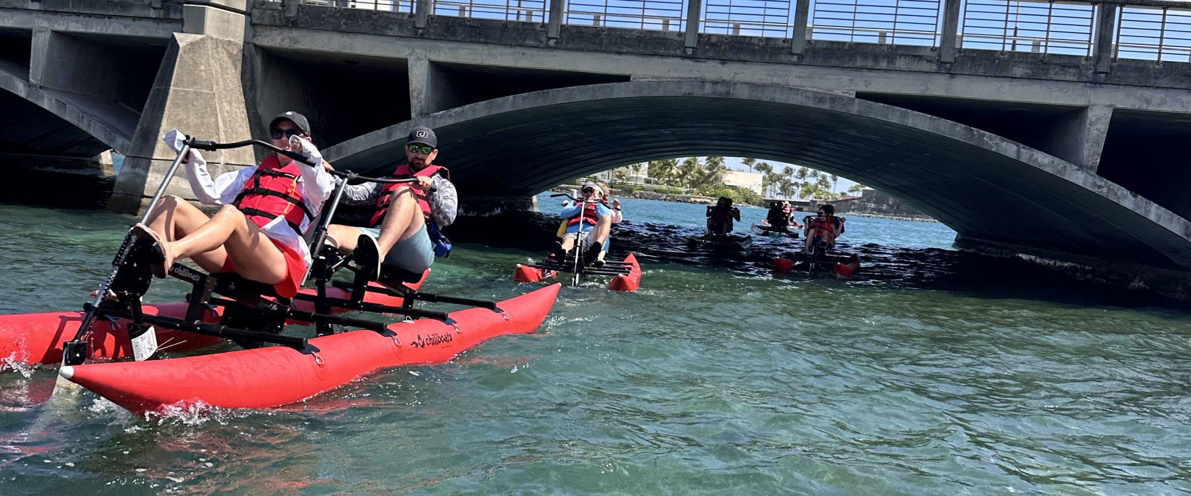 Two people ride a red waterbike on clear blue water near a historic fort on a sunny day.