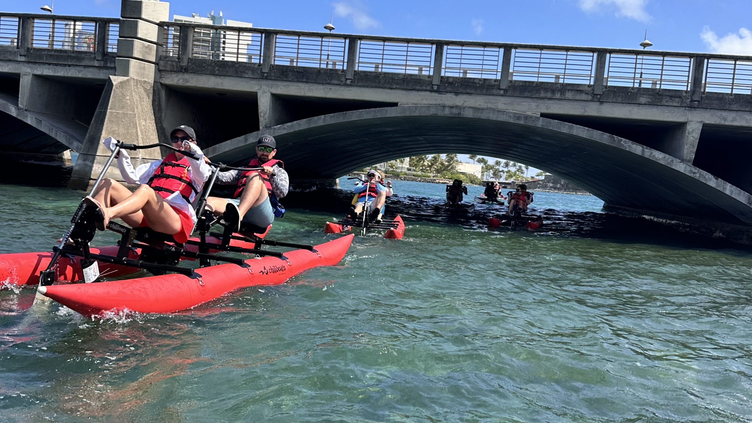 Two people ride a red waterbike on clear blue water near a historic fort on a sunny day.