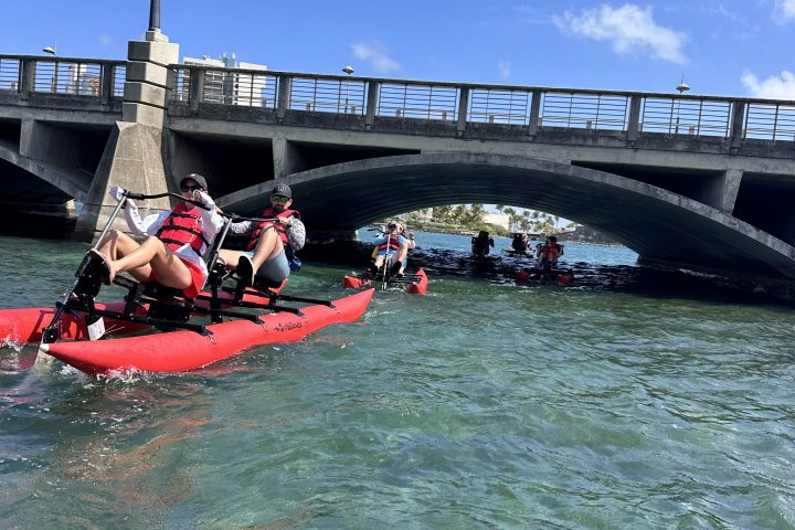 Two people ride a red waterbike on clear blue water near a historic fort on a sunny day.